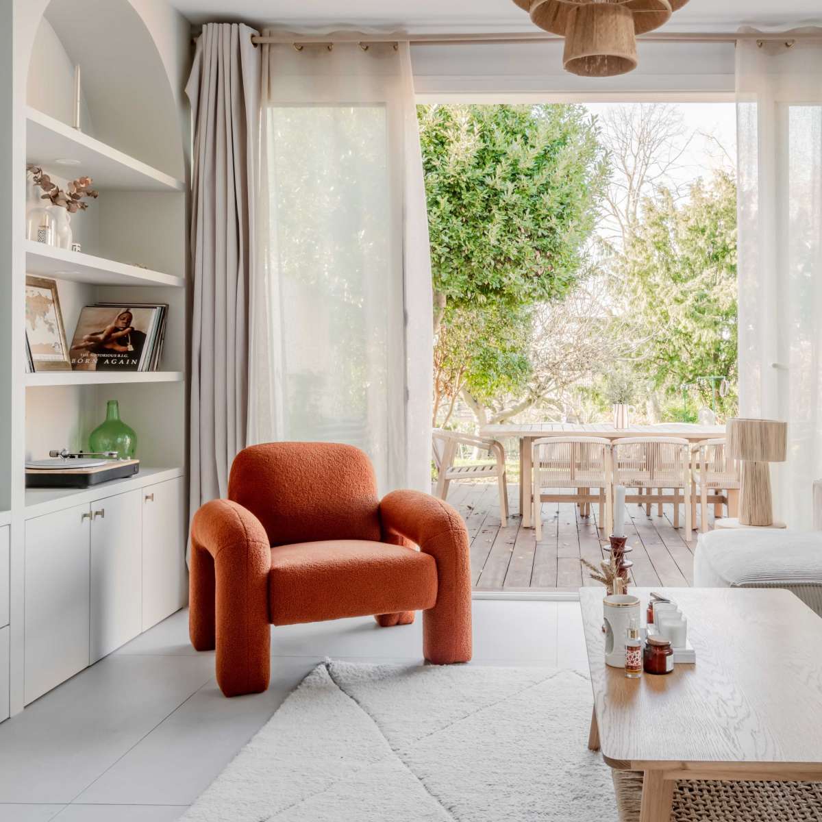 A cozy living room features a neutral sofa and an orange chair, with large windows that open to a wooden deck and a garden area.