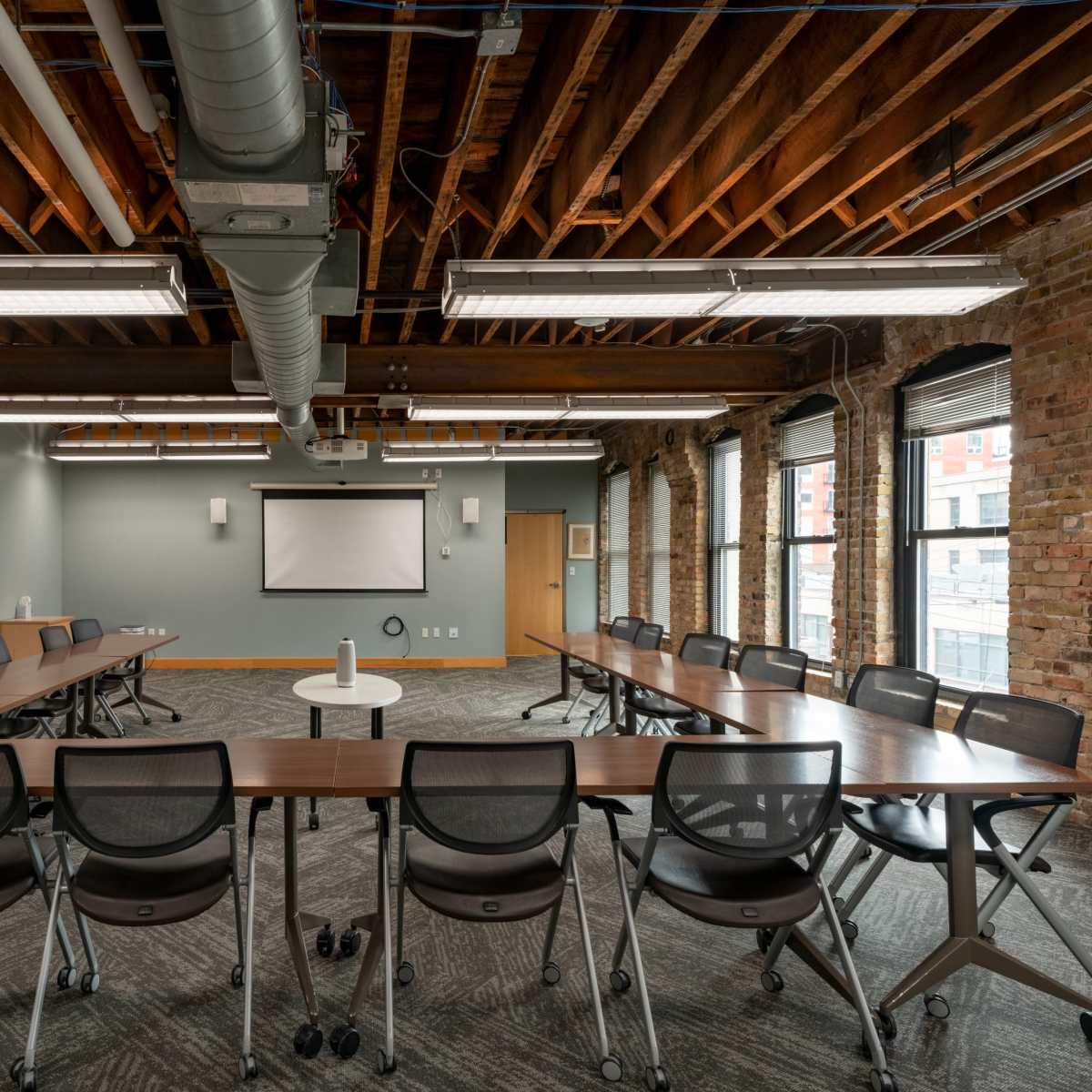 A spacious conference room with a large rectangular table surrounded by rolling chairs, exposed wooden beams, and large windows.