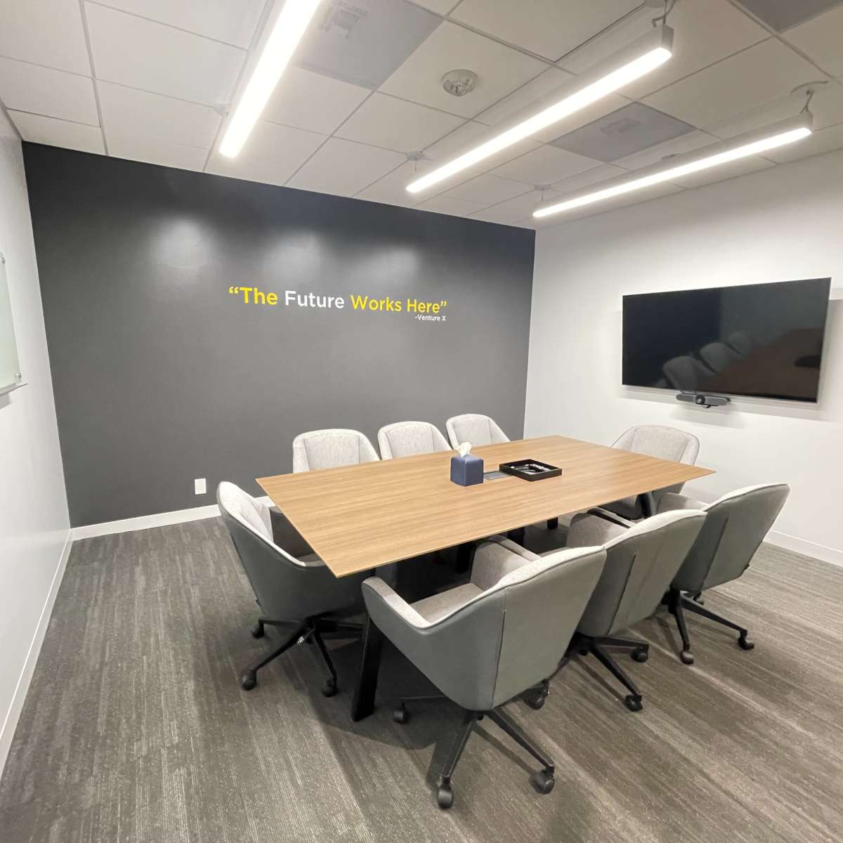 A modern conference room features a large wooden table surrounded by six upholstered chairs, with a wall-mounted screen and a motivational quote displayed above.