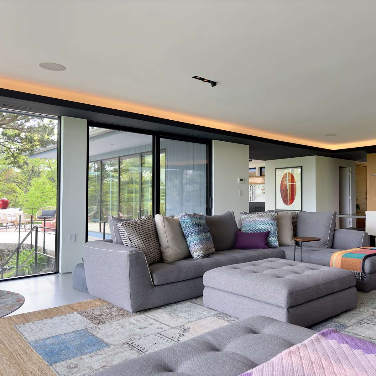 A modern living room with grey sectional sofas, a large area rug, and a dining area visible in the background.
