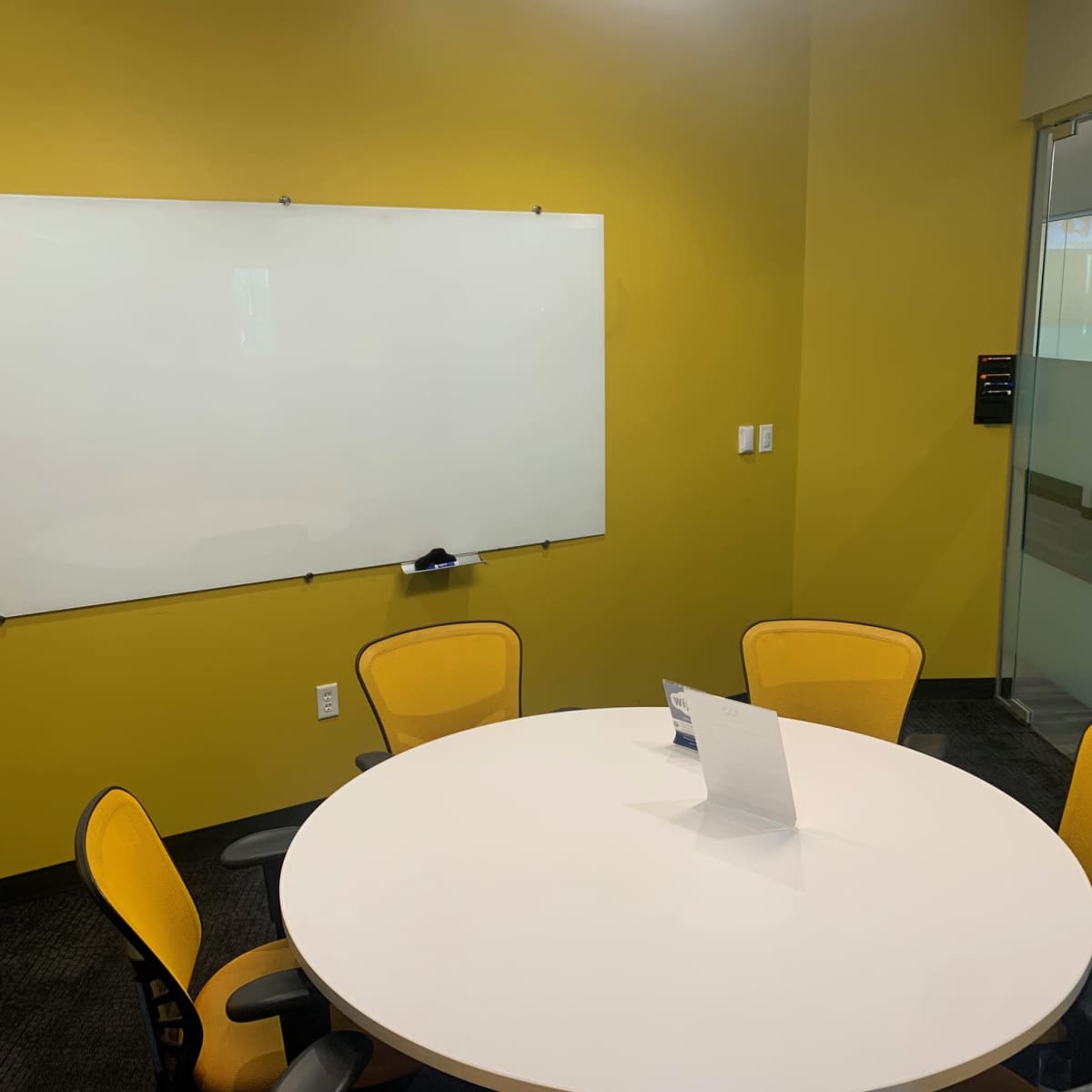 A small meeting room features a whiteboard, a round table with five yellow chairs, and a glass wall.