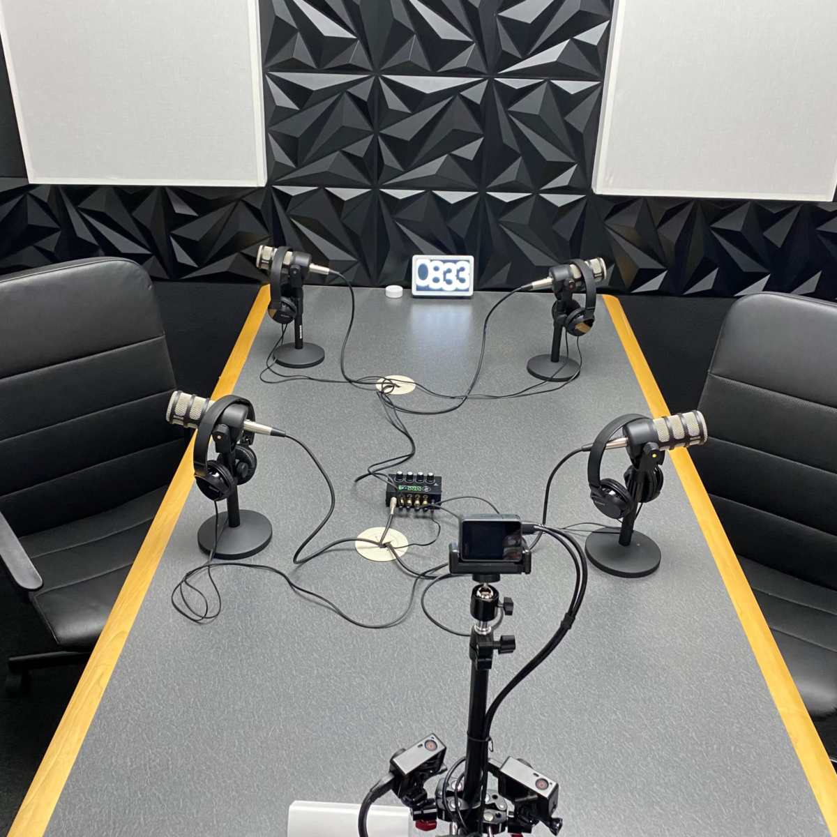 A podcast recording studio features a rectangular table with four microphones, two chairs, and a device for audio mixing, set against a textured black wall.