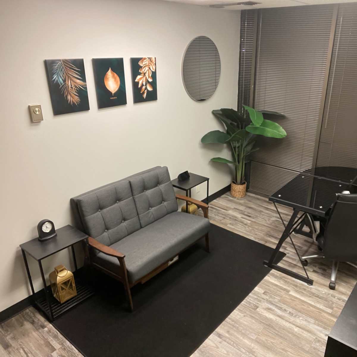 A small office space featuring a gray sofa, a glass desk, and a potted plant, complemented by framed artwork on the wall.