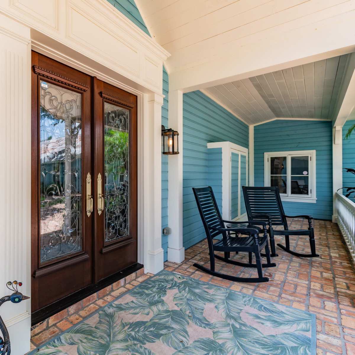 A front porch with blue wooden siding, featuring double doors with decorative glass, a patterned rug, and two black rocking chairs.