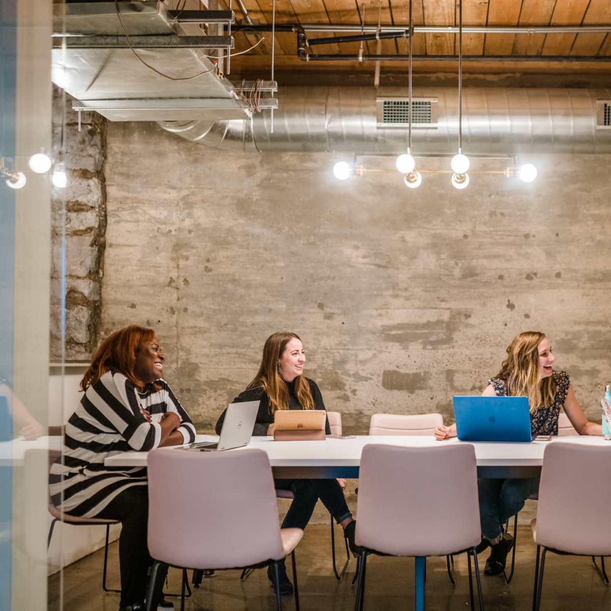 A group of five women is seated around a table in a modern meeting room with a concrete wall and overhead lighting.