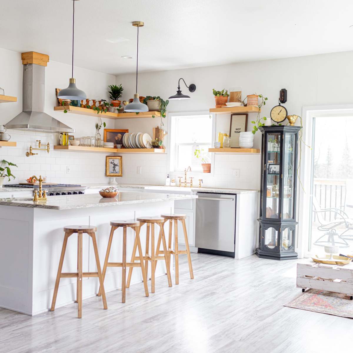 A modern kitchen with white cabinetry, a large island with wooden stools, and a mix of decorative plants and utensils on the shelves.