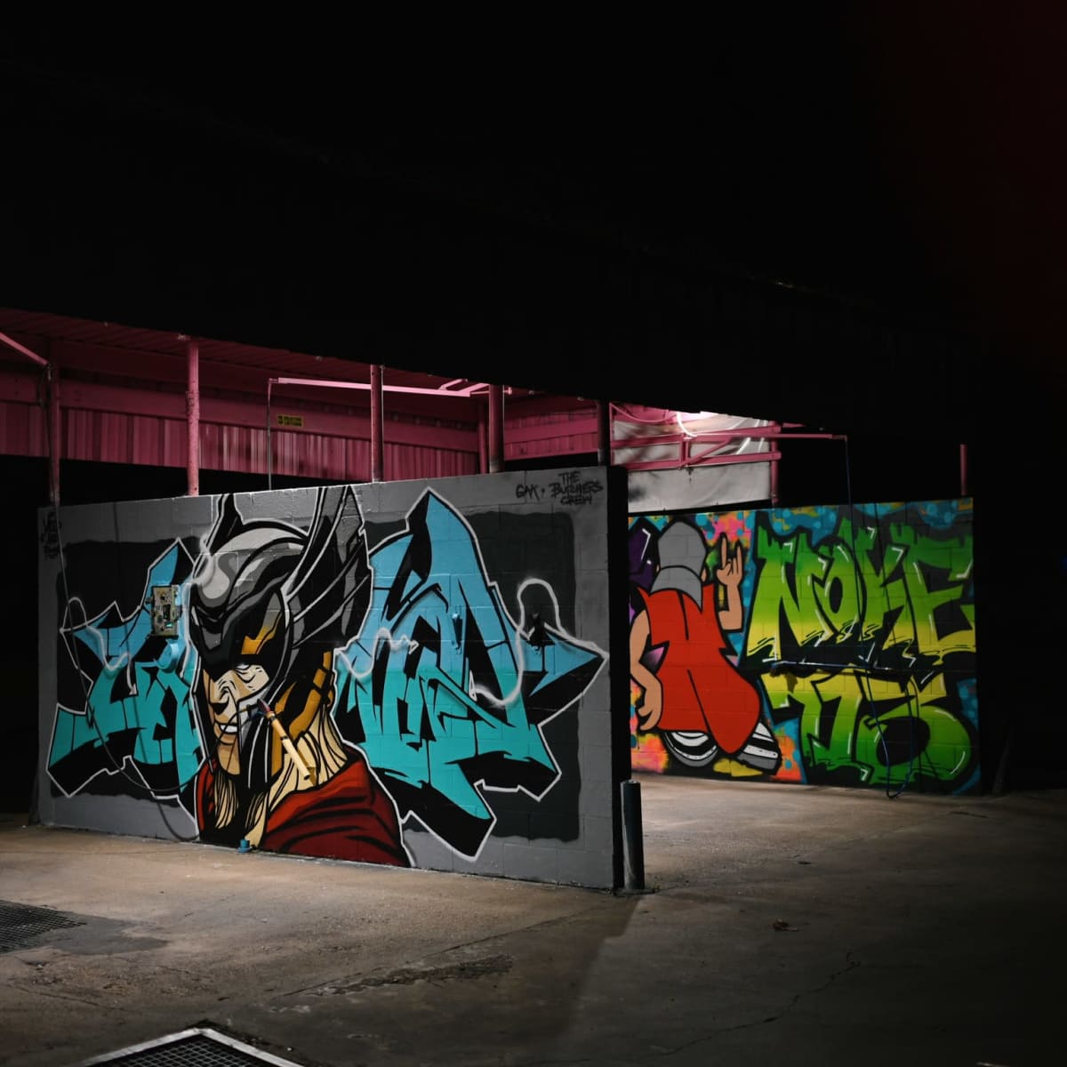Two vibrant murals painted on adjacent walls in a dimly lit industrial space at night.