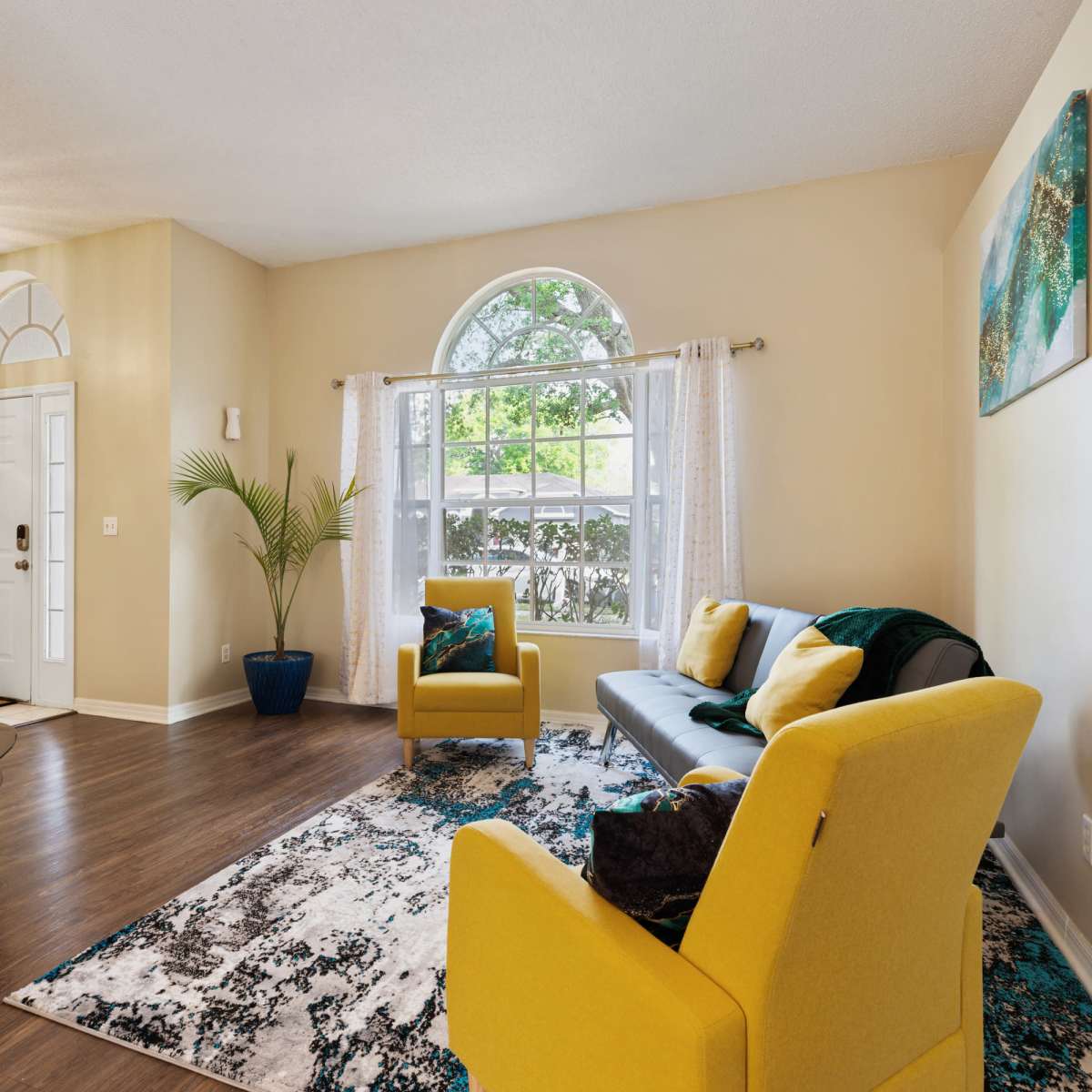 A living room features a gray sofa, two yellow chairs, and a large window with white curtains, surrounded by decorative artwork and a potted plant.