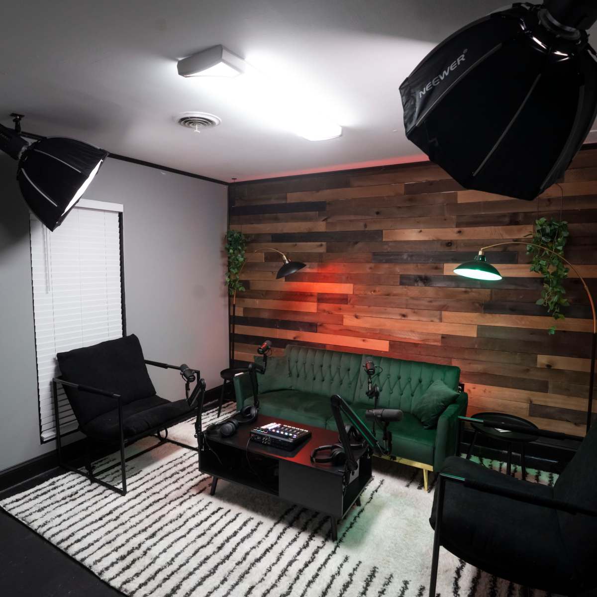 A recording studio features a green velvet couch, black chairs, a coffee table with equipment, and two large lights on tripods against a wooden accent wall.