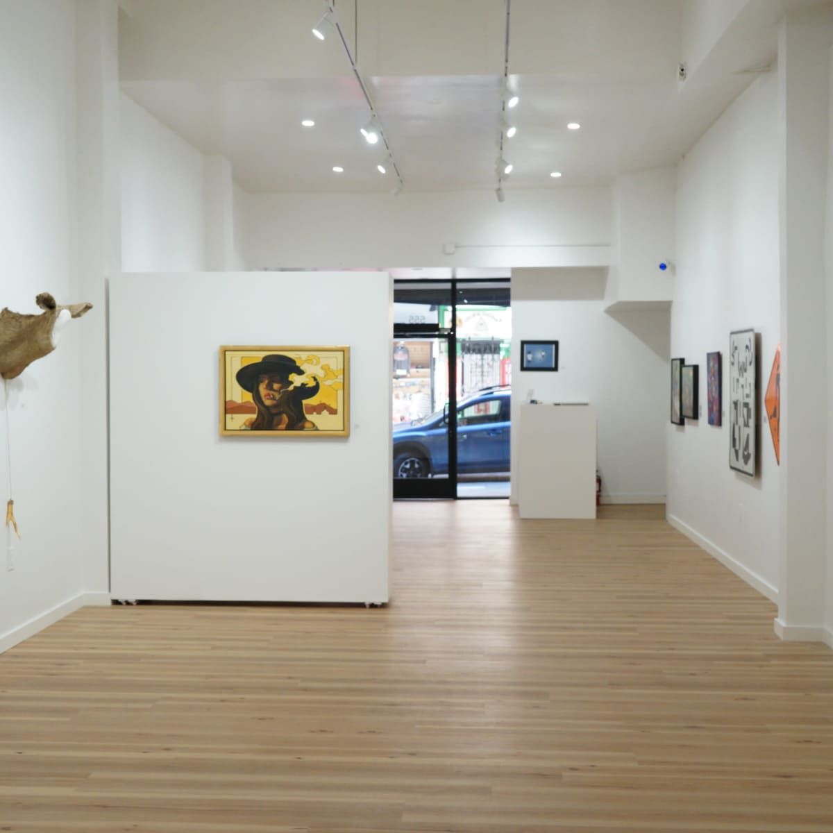 The interior of an art gallery features white walls, wooden flooring, and various artworks displayed, with a large window showing a car parked outside.