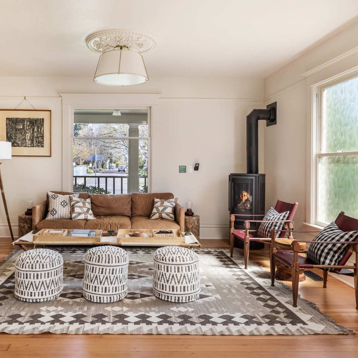 A cozy living room with a brown couch, a wooden coffee table, three patterned poufs, and a small wood stove beside large windows.