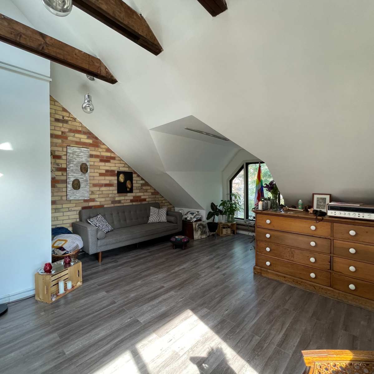 An attic room featuring a gray couch, a wooden dresser, and large windows with natural light illuminating the space.