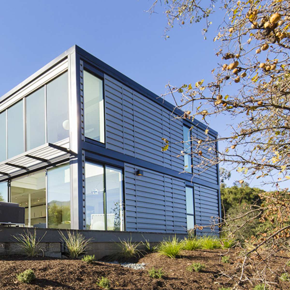 A modern, two-story house with a metal exterior and large windows is situated on a hillside surrounded by landscaping.