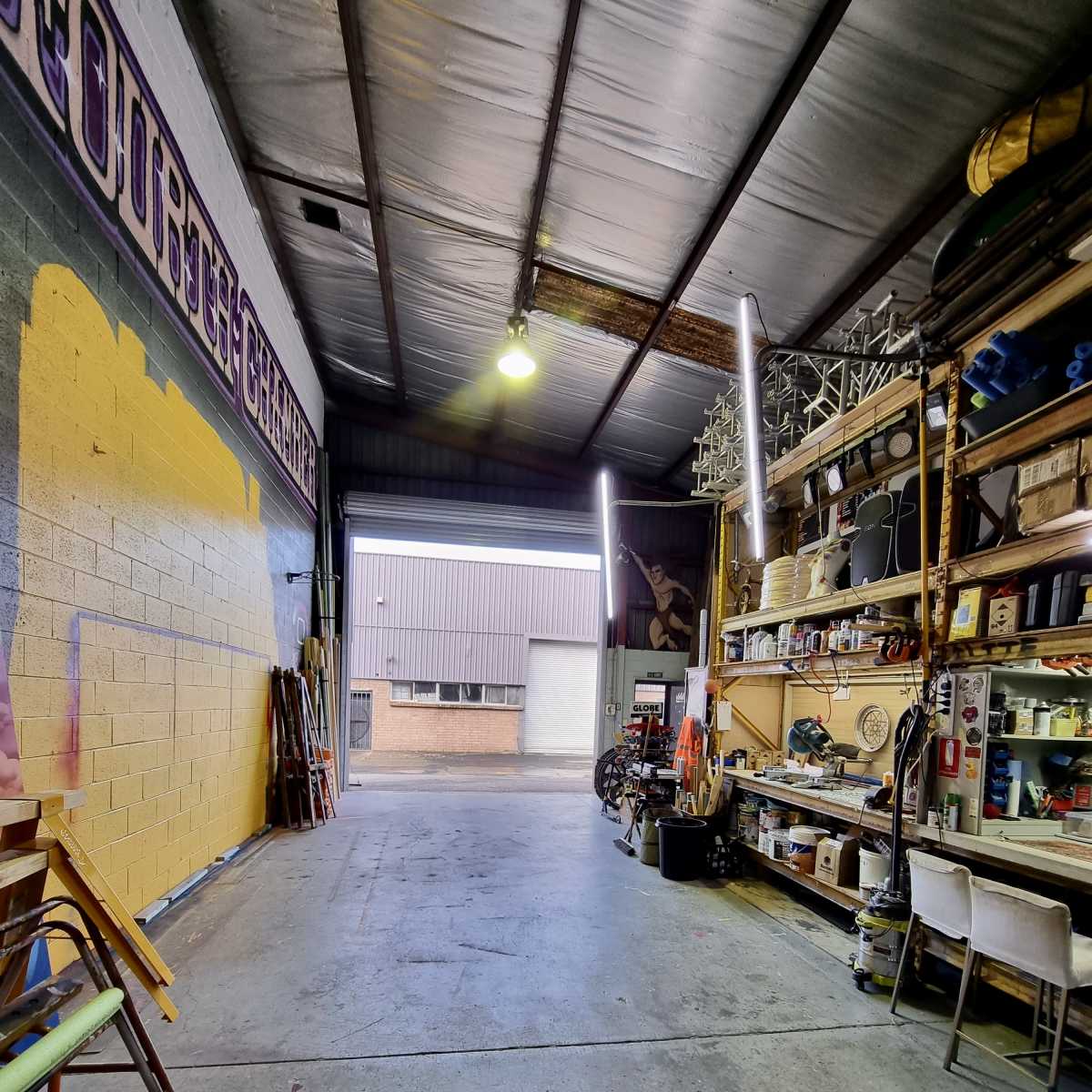 The interior of a spacious workshop with tools and materials organized on shelves along one side, illuminated by overhead lights.