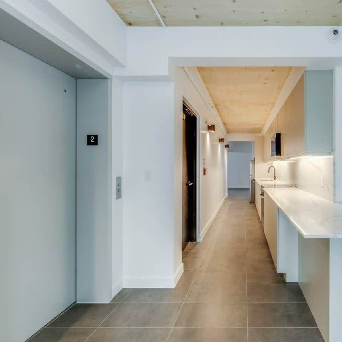 A modern hallway with an elevator on the left, a kitchen counter along the side, and doors leading to rooms at the end.