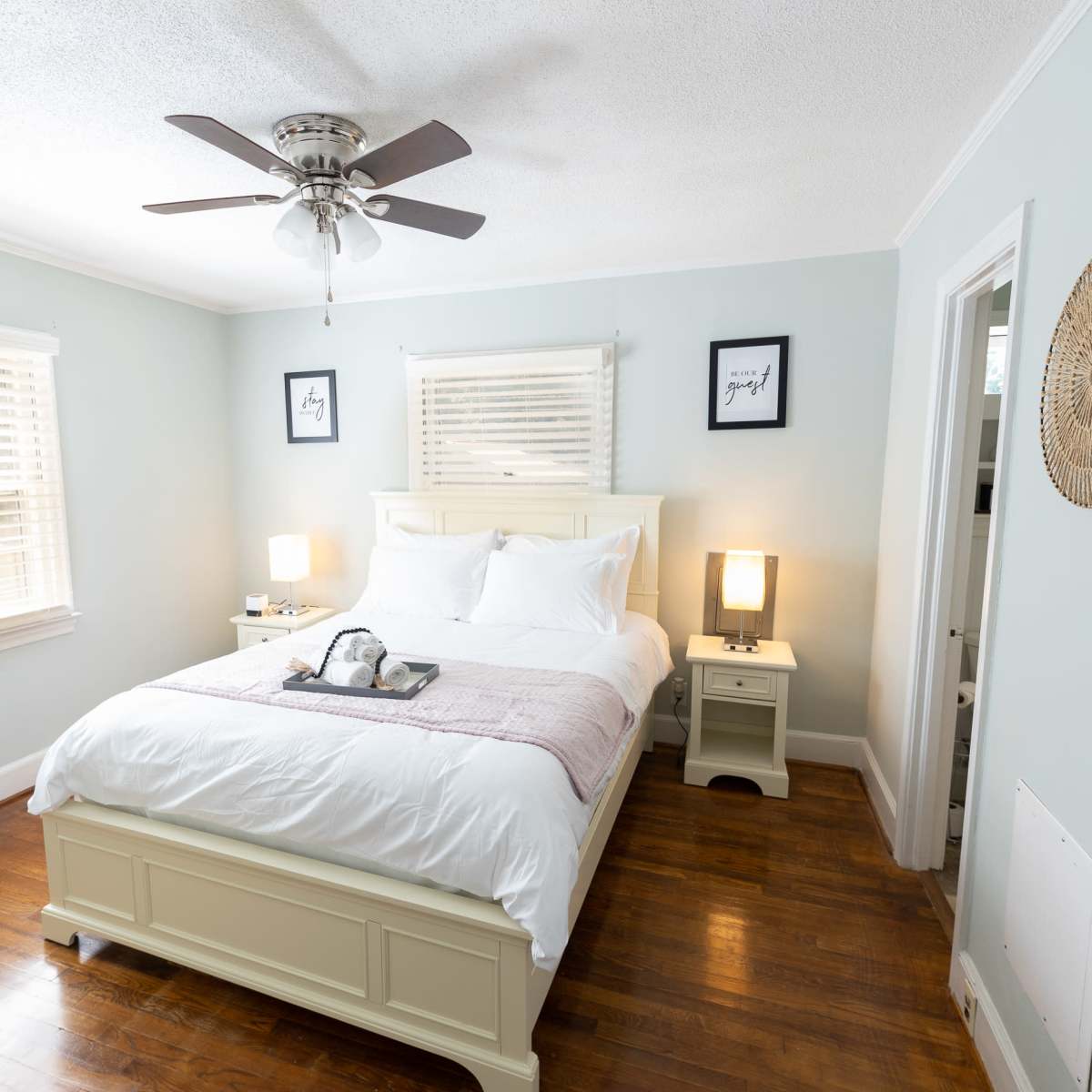 A well-lit bedroom features a double bed with white linens, a nightstand with a lamp, and a television in the corner.