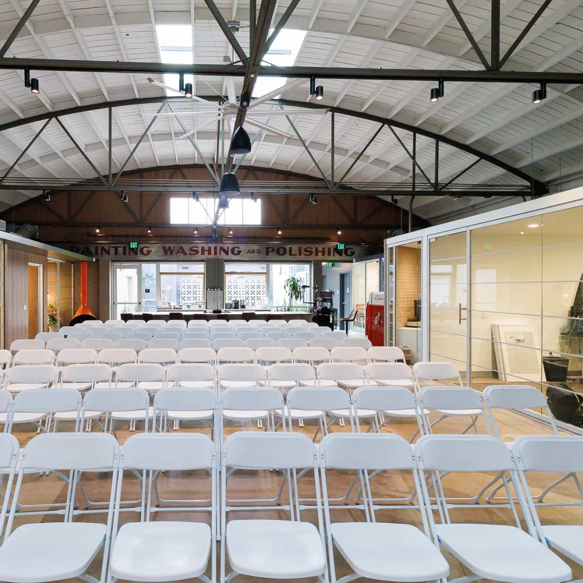 A spacious room with rows of white chairs facing a table in a well-lit area featuring large windows and high ceilings.