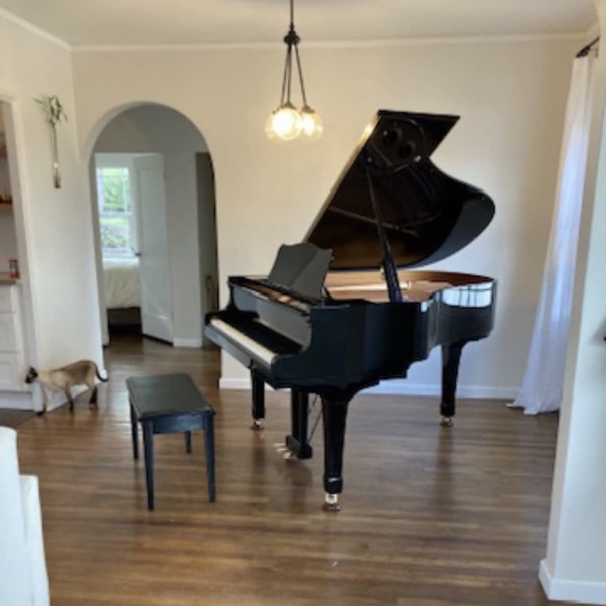A grand piano and a black bench are positioned in a well-lit room with wooden floors and an arched doorway in the background.