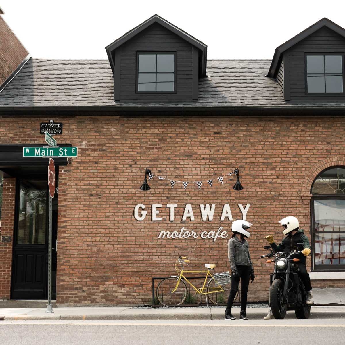 A woman in a helmet chats with a man in a motorcycle helmet outside the Getaway Motor Cafe, which features a brick building and decorative signage.