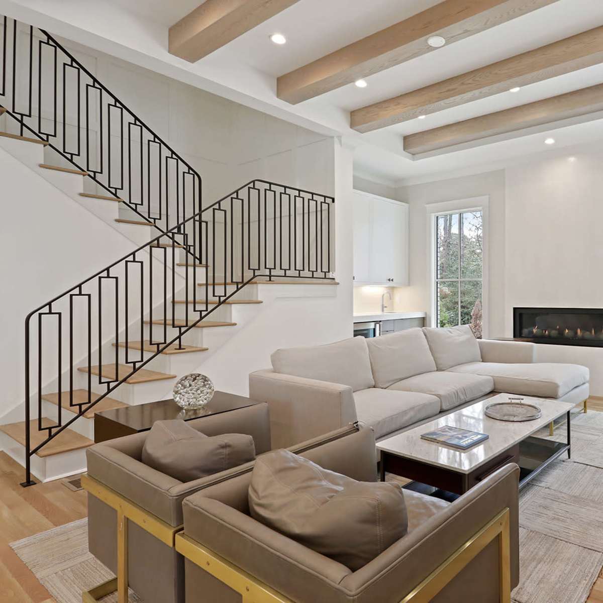 A modern living room with a sectional sofa, a coffee table, and a staircase featuring wrought iron railings, illuminated by natural light from large windows.