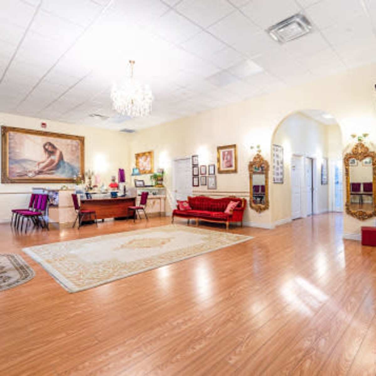 A spacious room with wooden flooring, featuring a chandelier, a large painting, various seating areas, and multiple doorways leading to adjacent rooms.