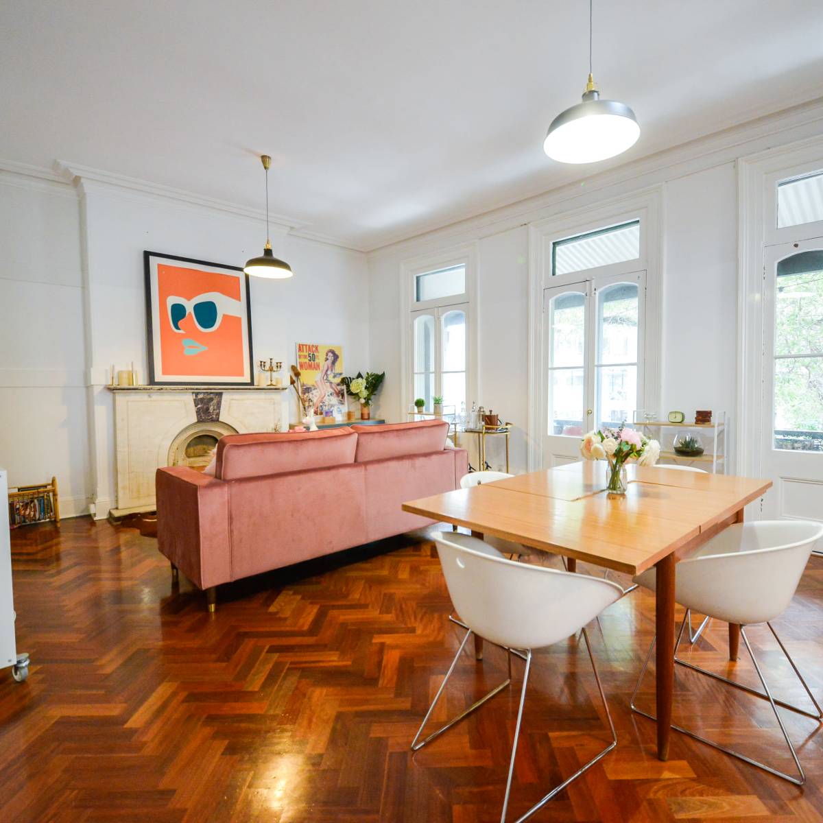 A modern living space features a pink sofa, a wooden dining table with white chairs, and large windows leading to a balcony.