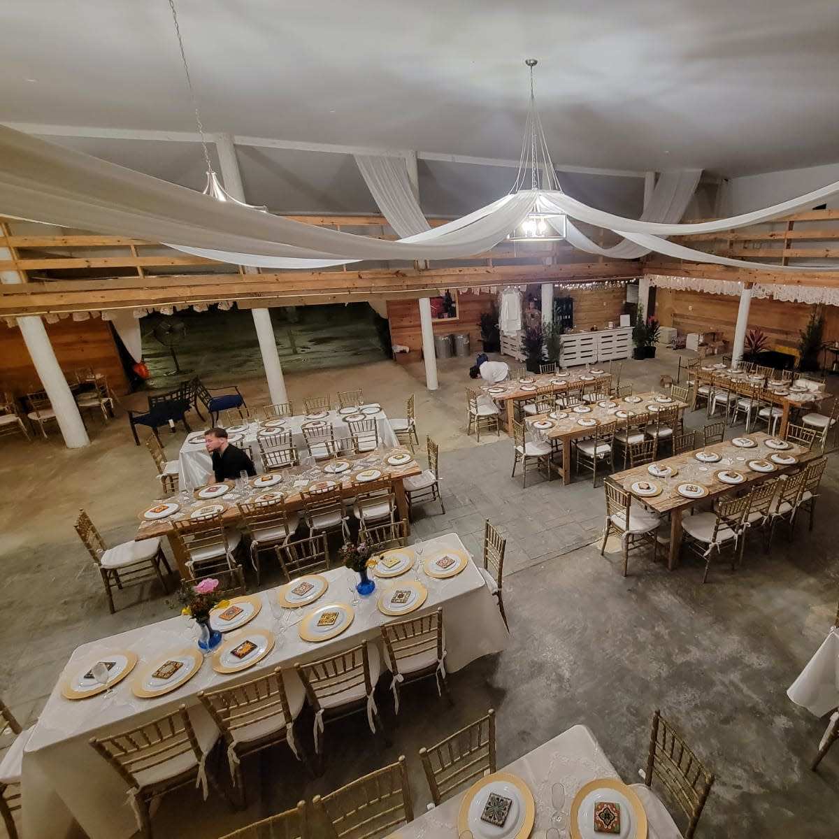 A spacious event hall set with multiple tables arranged for a dining event, featuring gold-colored chairs and table settings.