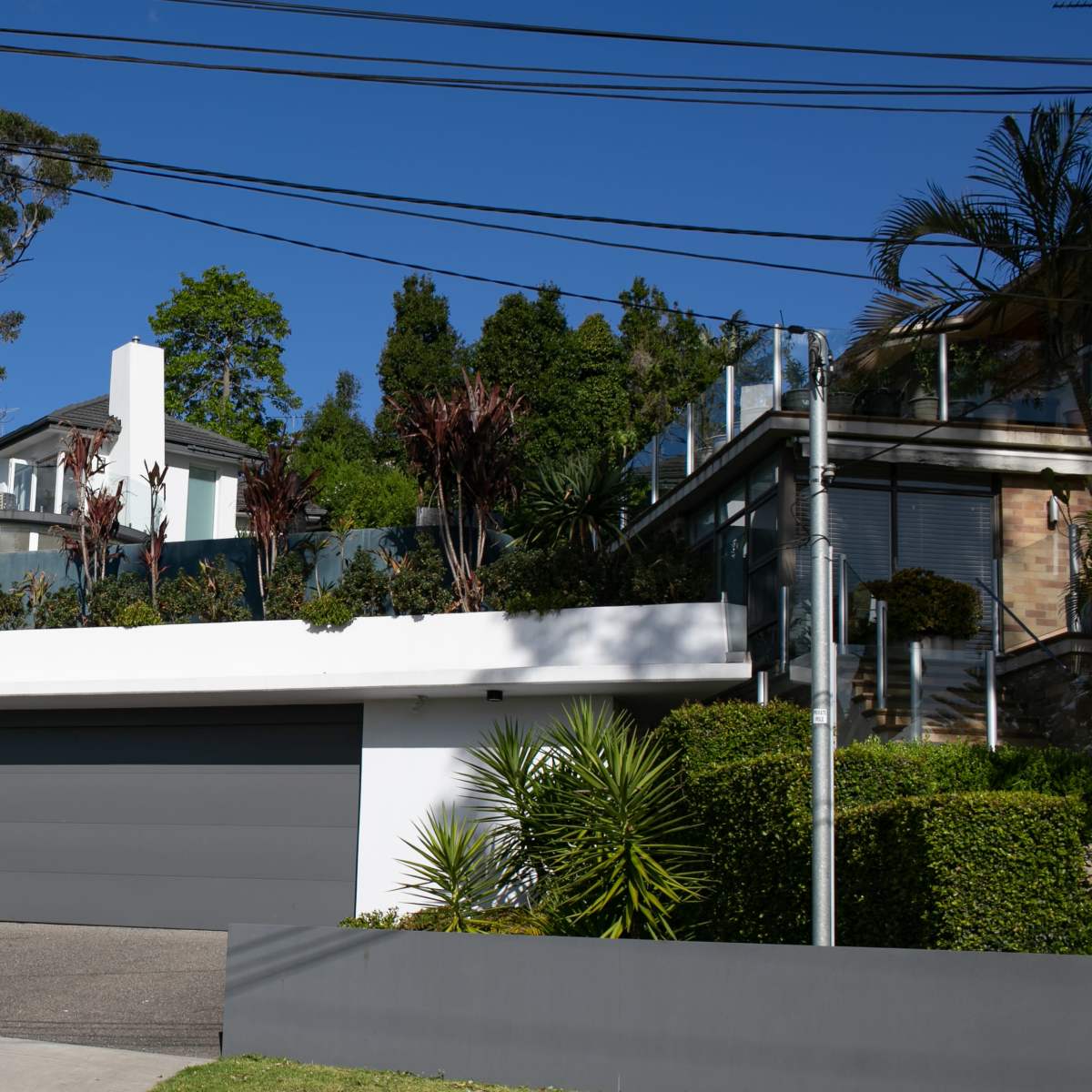 A house with a flat green roof and a gray garage door is situated on a sloped lot surrounded by trees and plants.