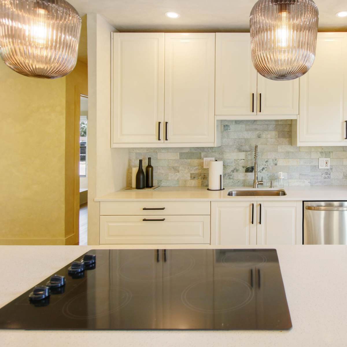 A modern kitchen features white cabinets, a stainless steel sink, and a smooth countertop with a built-in cooktop, illuminated by pendant lights.