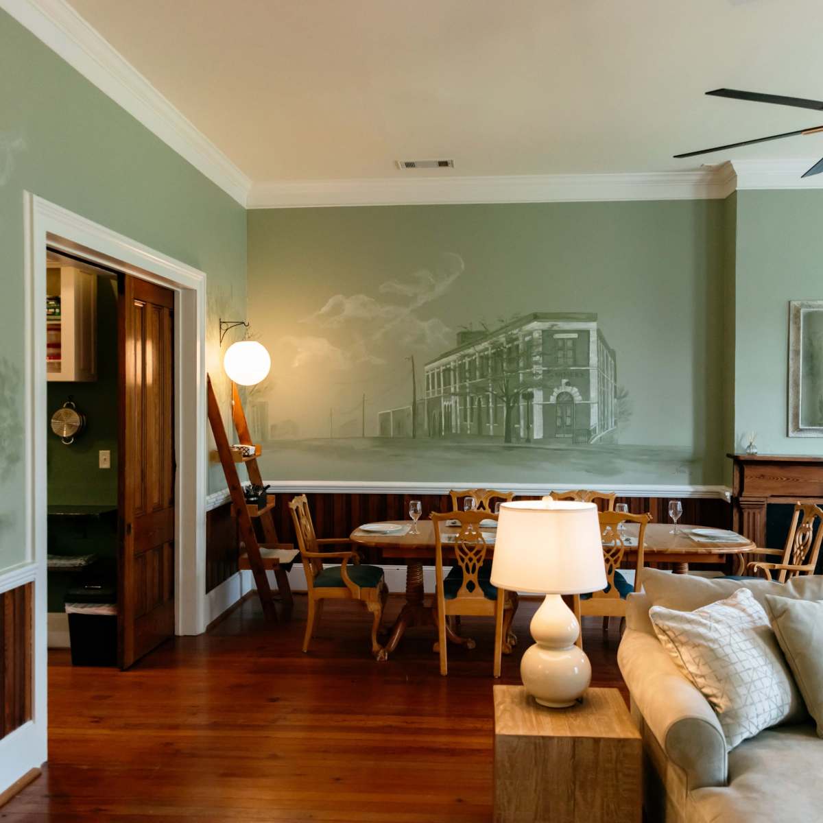 The room features green walls with a mural of a historical building, a dining area with wooden chairs, and a living space with a sofa and lamp.
