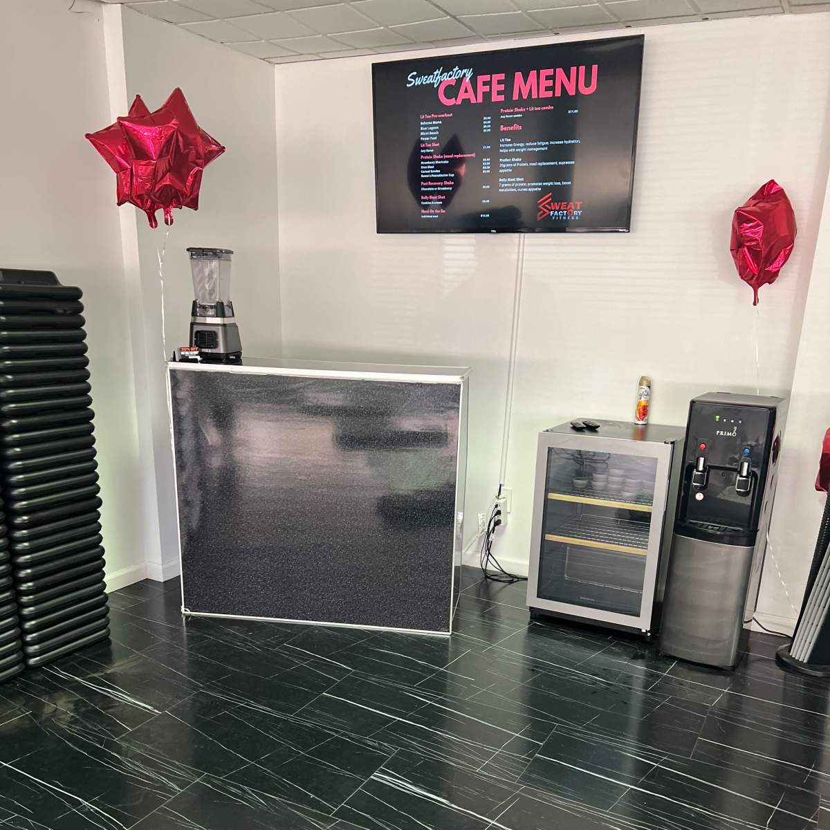 A cafe area featuring a counter with a blender, a menu display on the wall, a beverage cooler, a water dispenser, and decorative red balloons.