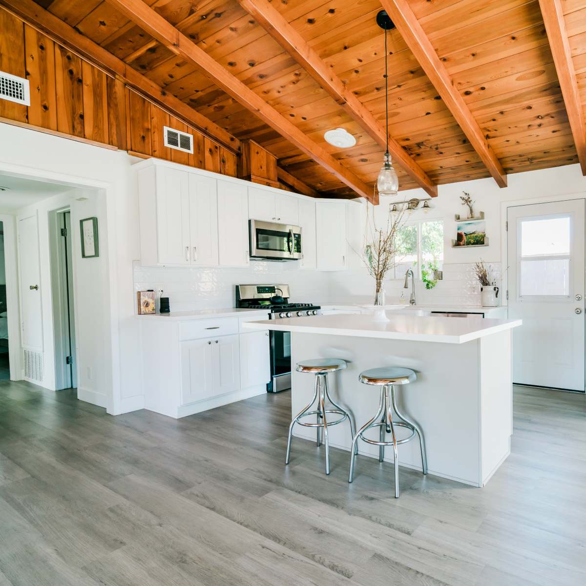 A modern kitchen features white cabinetry, stainless steel appliances, and a central island with bar stools, all beneath a wooden beamed ceiling.