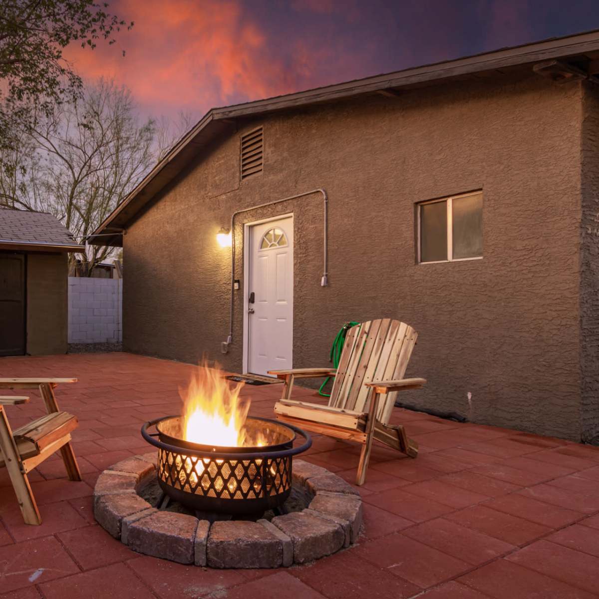 A circular fire pit with flames sits between two wooden lounge chairs on a brick patio, adjacent to a square building at dusk.