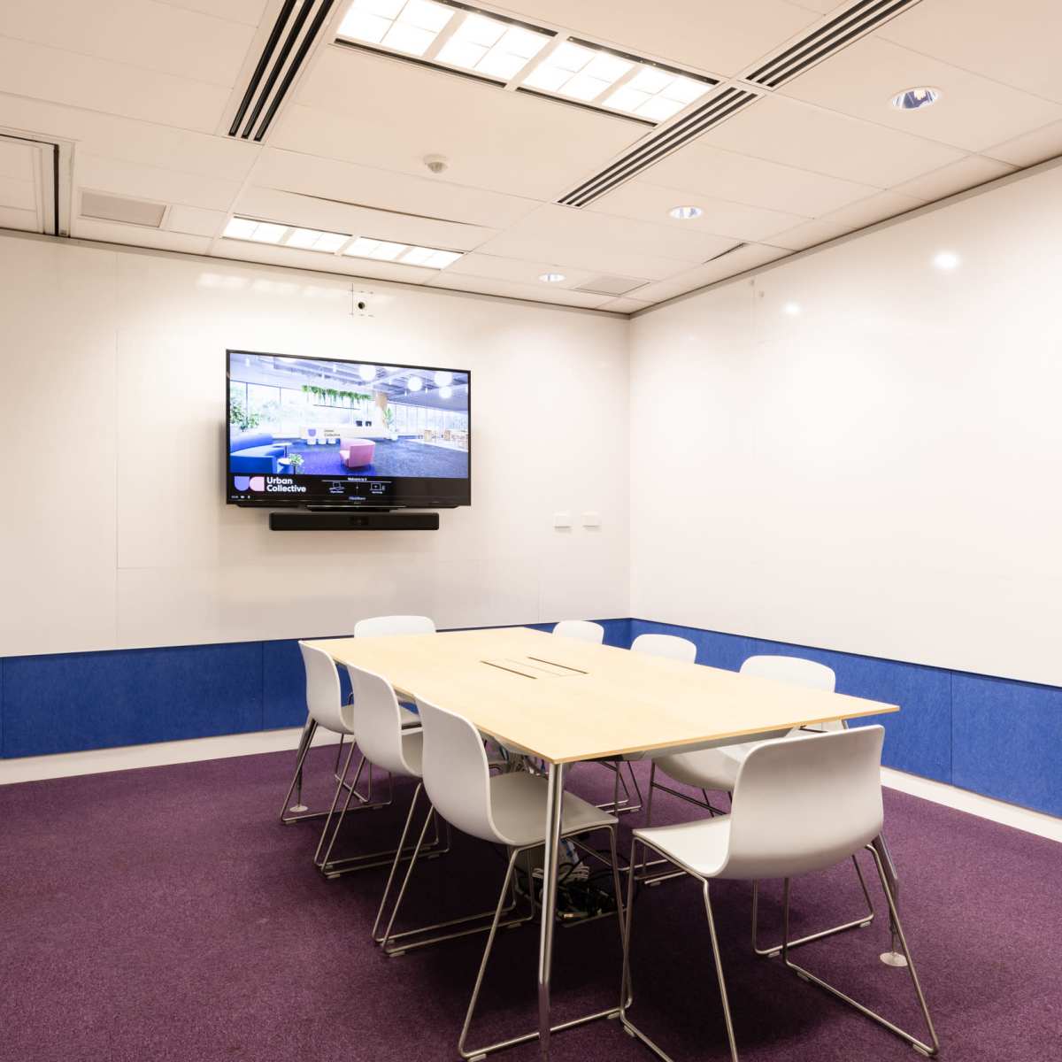 A modern conference room with a wooden table surrounded by white chairs and a wall-mounted television.