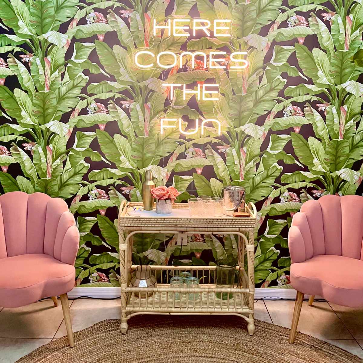 The scene features a brightly decorated wall with tropical leaf wallpaper, a glowing neon sign that reads "HERE COMES THE FUN," and two pink chairs beside a vintage-looking table adorned with a small vase and a metal cup.