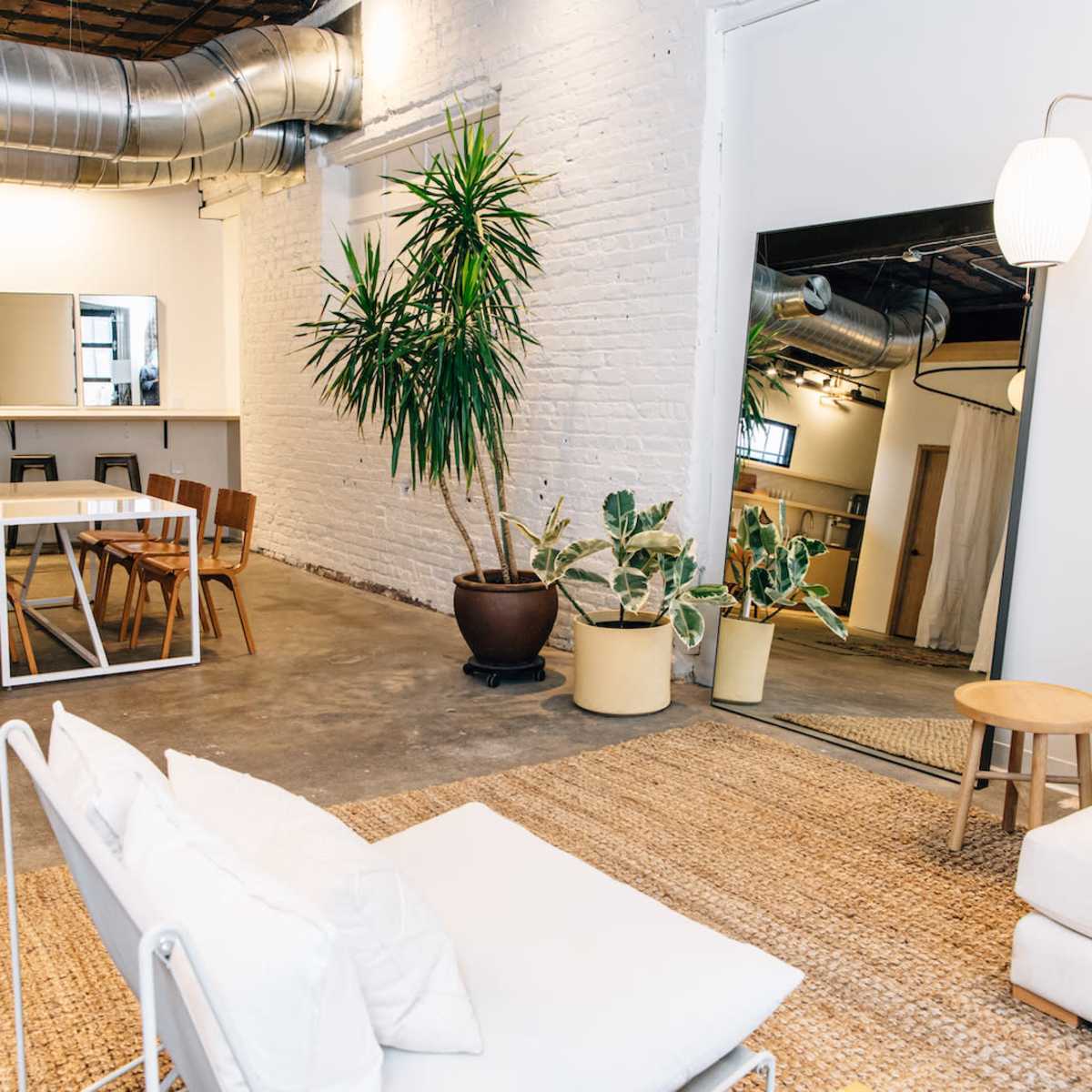 A spacious, industrial-style room featuring a seating area with a white sofa, a dining table, wooden chairs, and various potted plants.