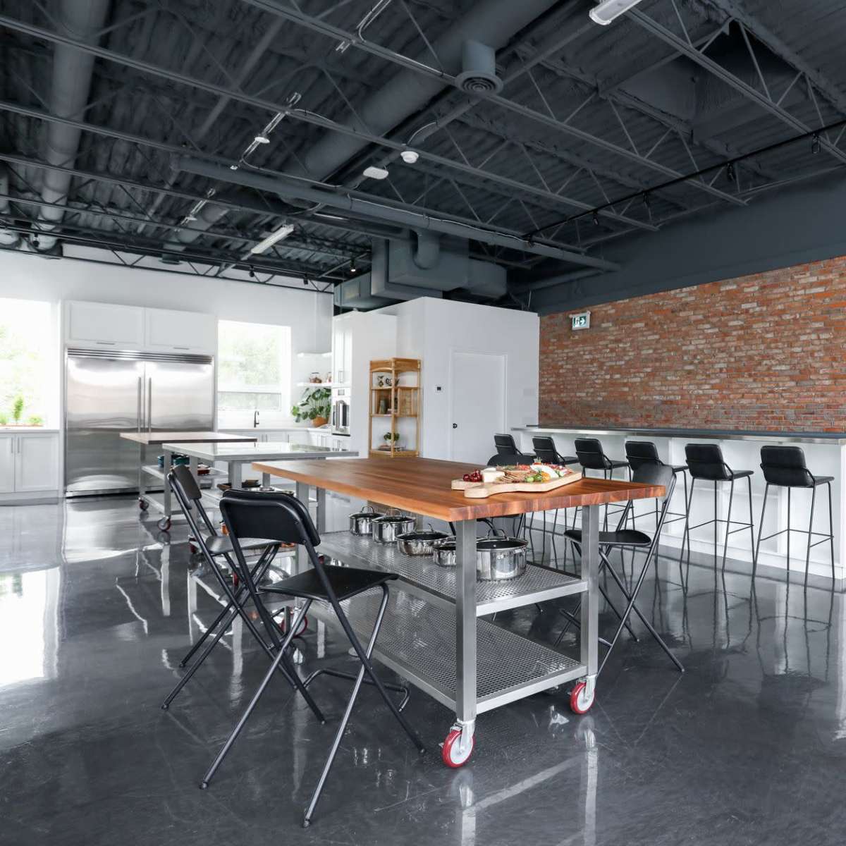 A modern kitchen with a large wooden island, metal stools, and a brick wall, featuring stainless steel appliances and a minimalistic design.