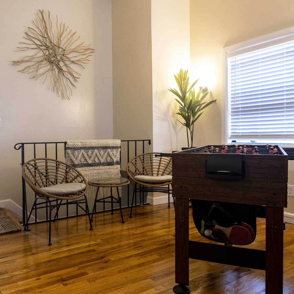 A room with a foosball table in the foreground, two wicker chairs and a decorative wall piece, with a doorway leading to another room in the background.