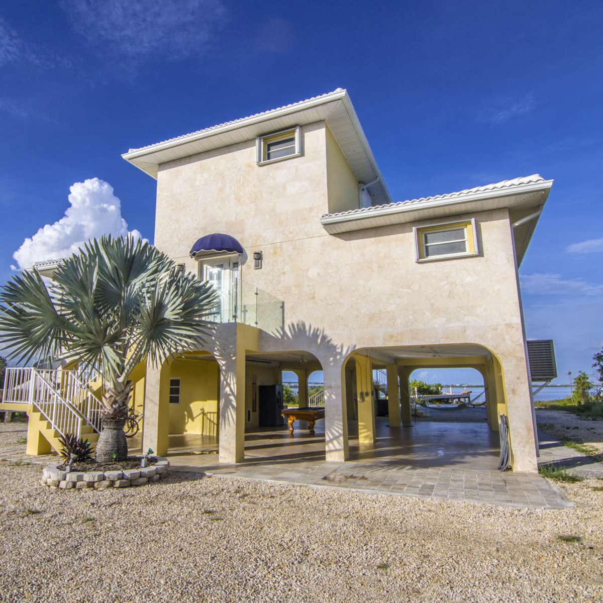 A two-story beige house with a balcony and open spaces underneath is surrounded by gravel, palm trees, and a clear blue sky.
