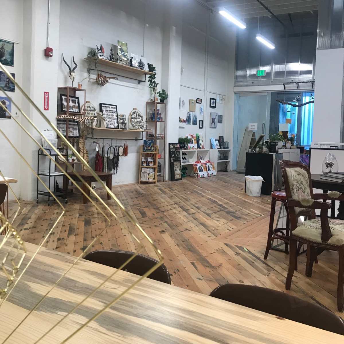 A spacious, brightly lit indoor area featuring wooden flooring, shelves with various decorative items, and a few tables and chairs arranged throughout.