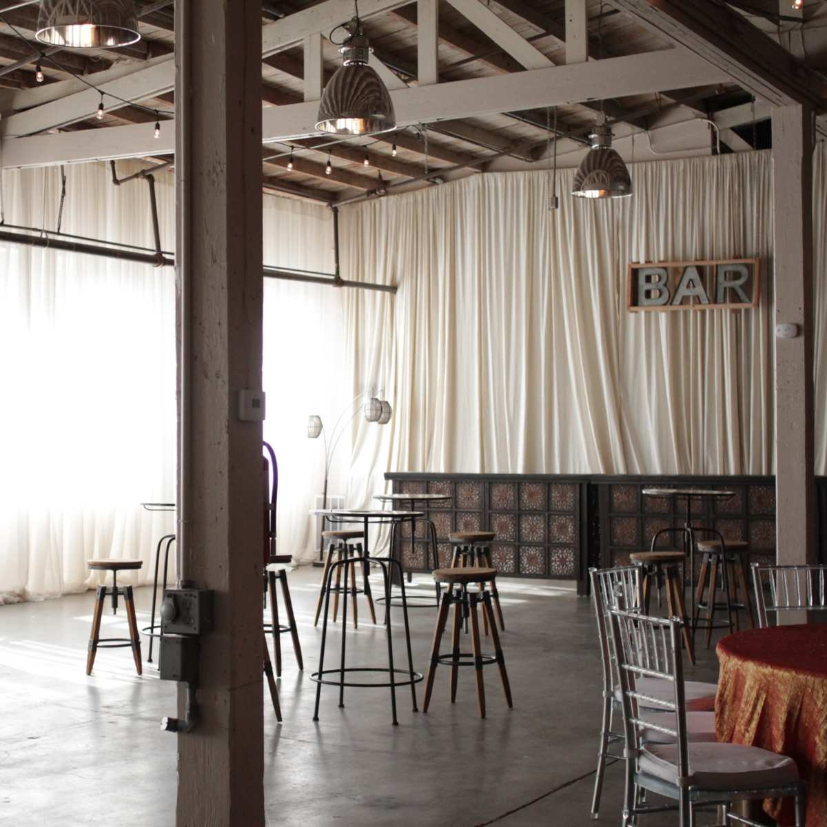 An indoor venue with tall bar tables and stools arranged among large windows draped with sheer curtains, alongside a bar area.