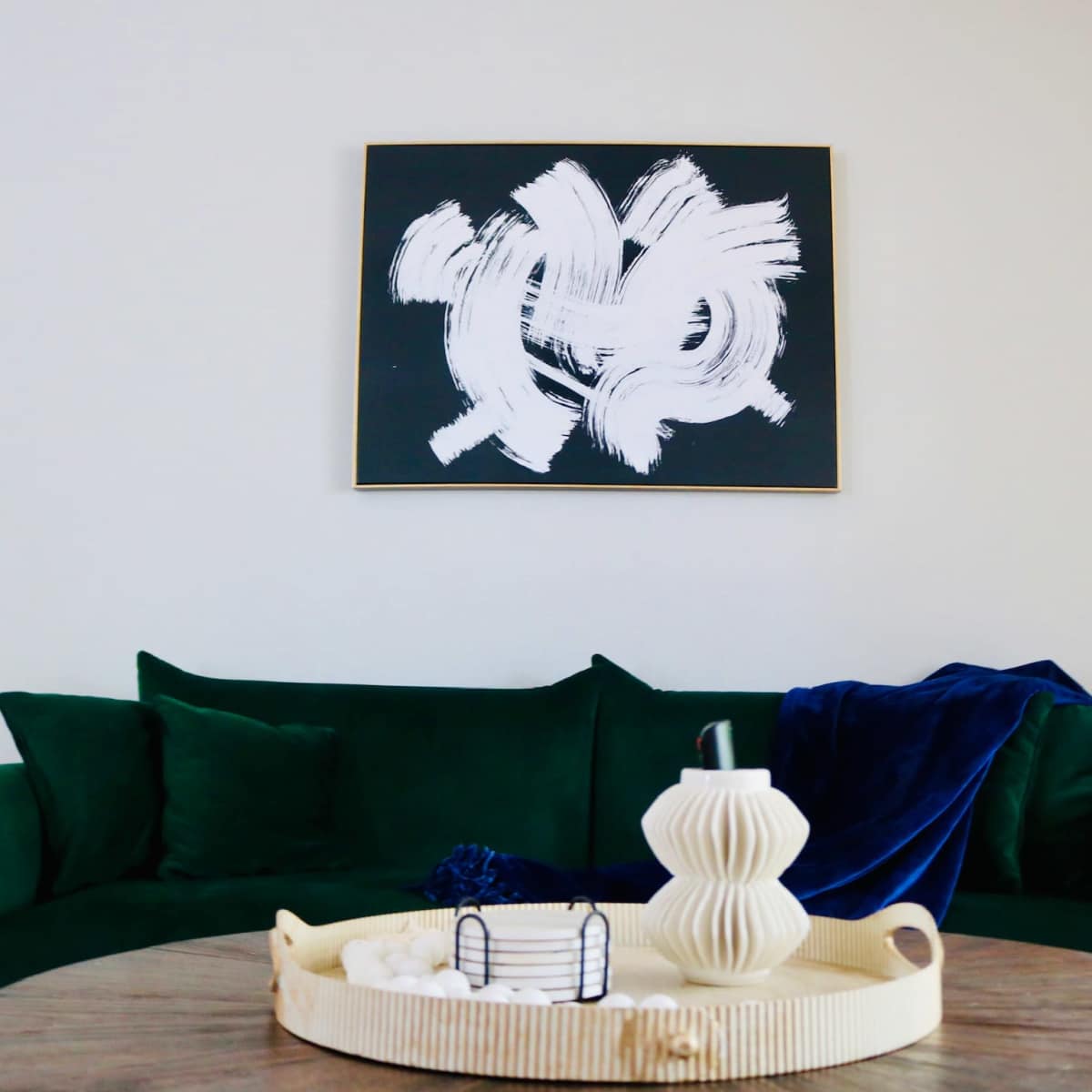 A green sofa is positioned in front of a black and white abstract painting, with a round wooden table topped with decorative items.