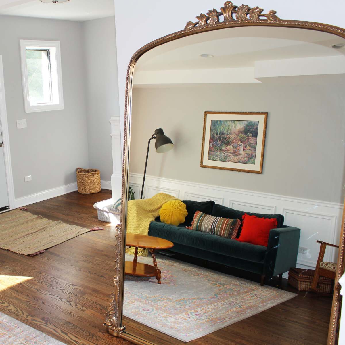 A living room with a green sofa, two armchairs, a round wooden table, and a large mirror reflecting the space, featuring warm colors and wooden flooring.
