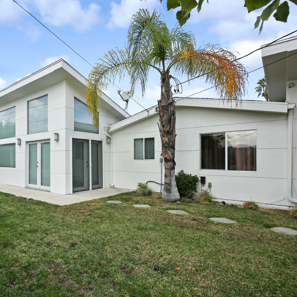 A modern two-story house with large windows, a manicured lawn, and a palm tree in the backyard.