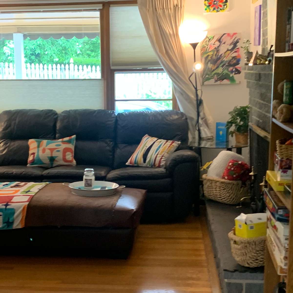 A living room featuring a black leather sofa with colorful throw pillows, a coffee table in front, and a bookshelf filled with various items.