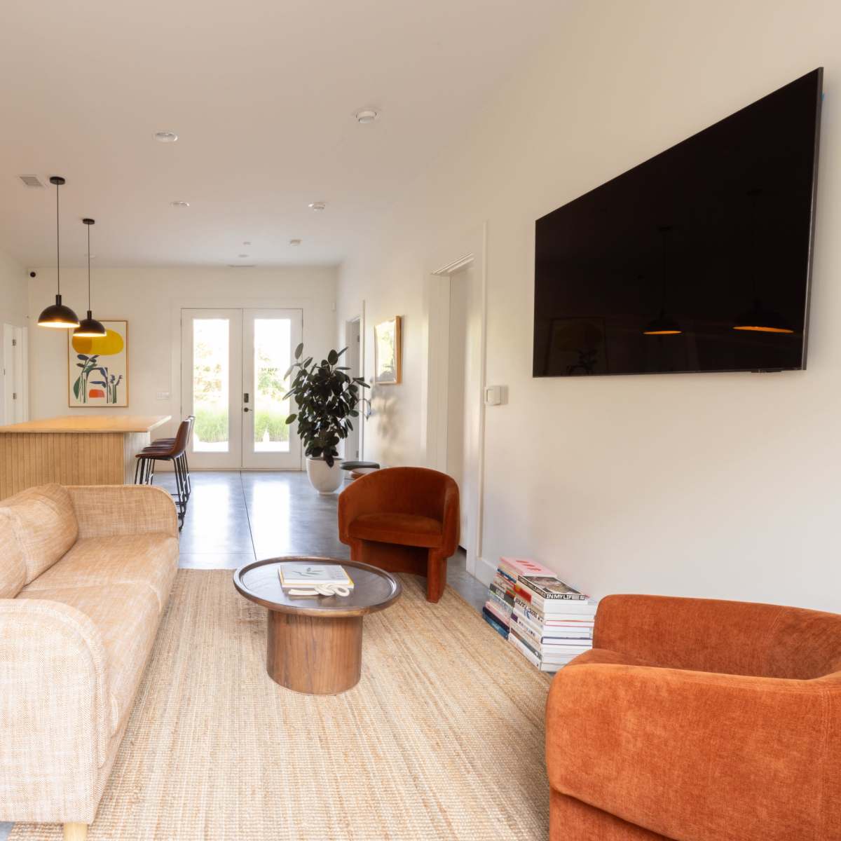 A modern living room features a beige sofa, two orange chairs, a round coffee table, and a television mounted on the wall, adjacent to a dining area with a wooden table and chairs.