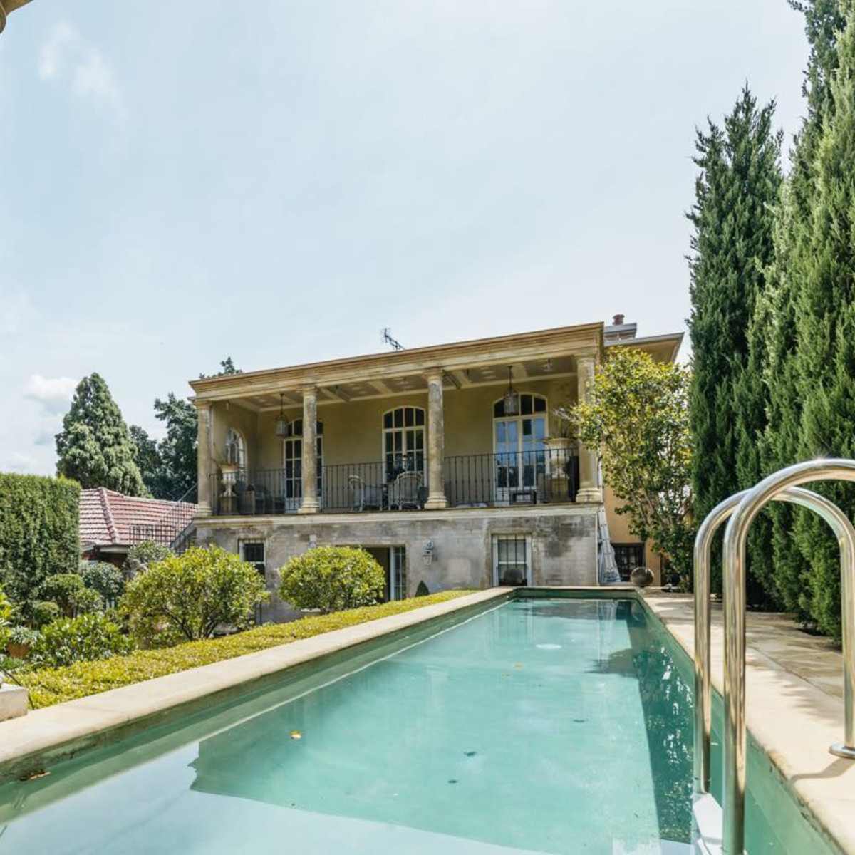 A modern house with a slim swimming pool, framed by manicured hedges and a stone pathway, under a clear sky.