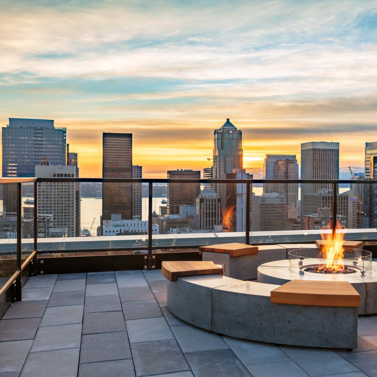 A circular concrete fire pit with wooden benches is surrounded by a rooftop terrace overlooking a city skyline at sunset.
