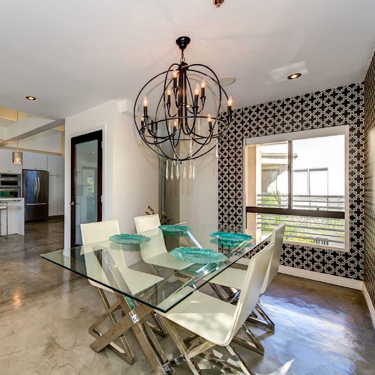 A modern dining area features a glass table with white chairs and a chandelier, adjacent to a kitchen with stainless steel appliances.