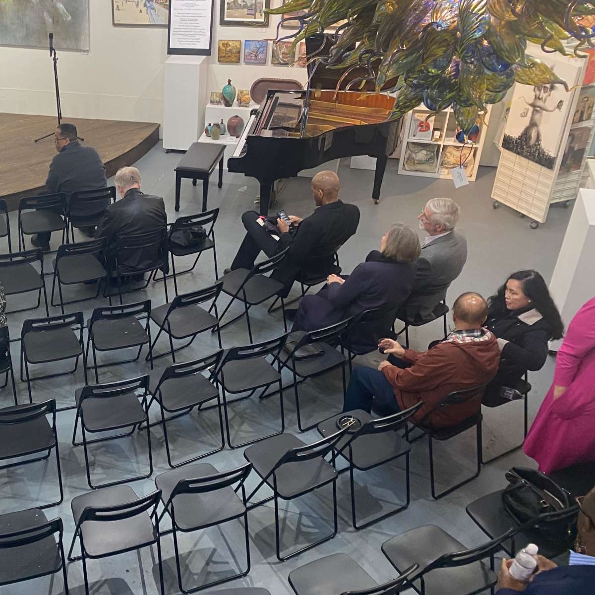 An indoor space set up for an event, with rows of empty black chairs facing a small stage, and attendees seated throughout the room.